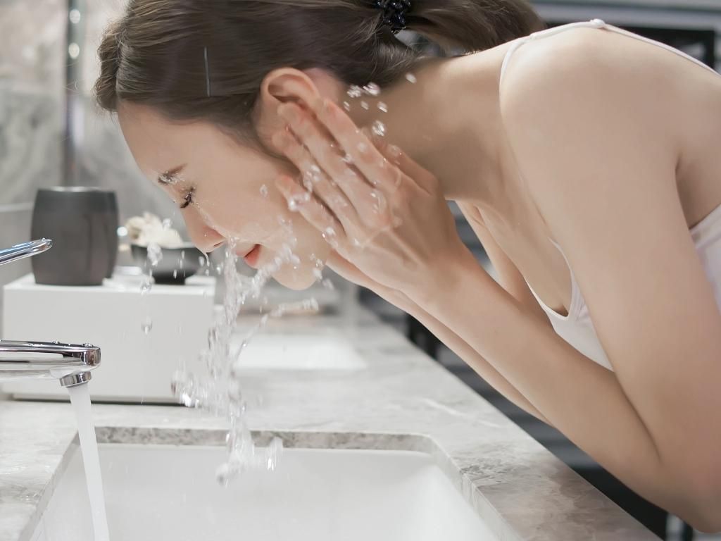 Retrato de una mujer joven que se lava la cara con agua y jabón natural mientras abre el grifo.
