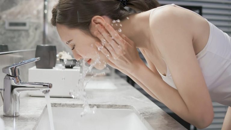 Foto de una mujer joven lavándose la cara con jabones naturales y agua mientas está el grifo abierto.
